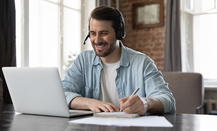 smiling man using laptop with headset taking notes while working from home five tips for productivity