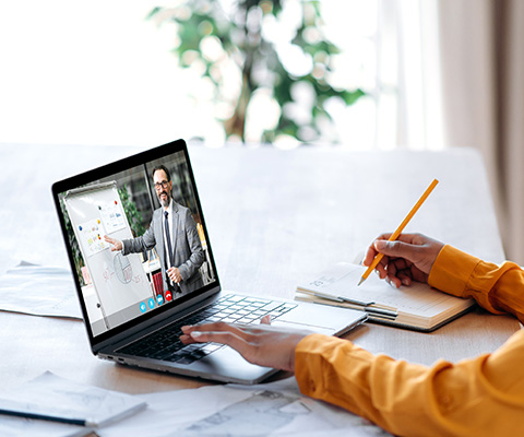 person attending a virtual meeting on a laptop while taking notes with a pencil on a desk surrounded by papers engaging in online collaboration in a modern workspace