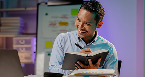 man writing in notebook with computer in a modern office setting smiling while engaged in work or study focused on learning and productivity 2 techniques 2 insights